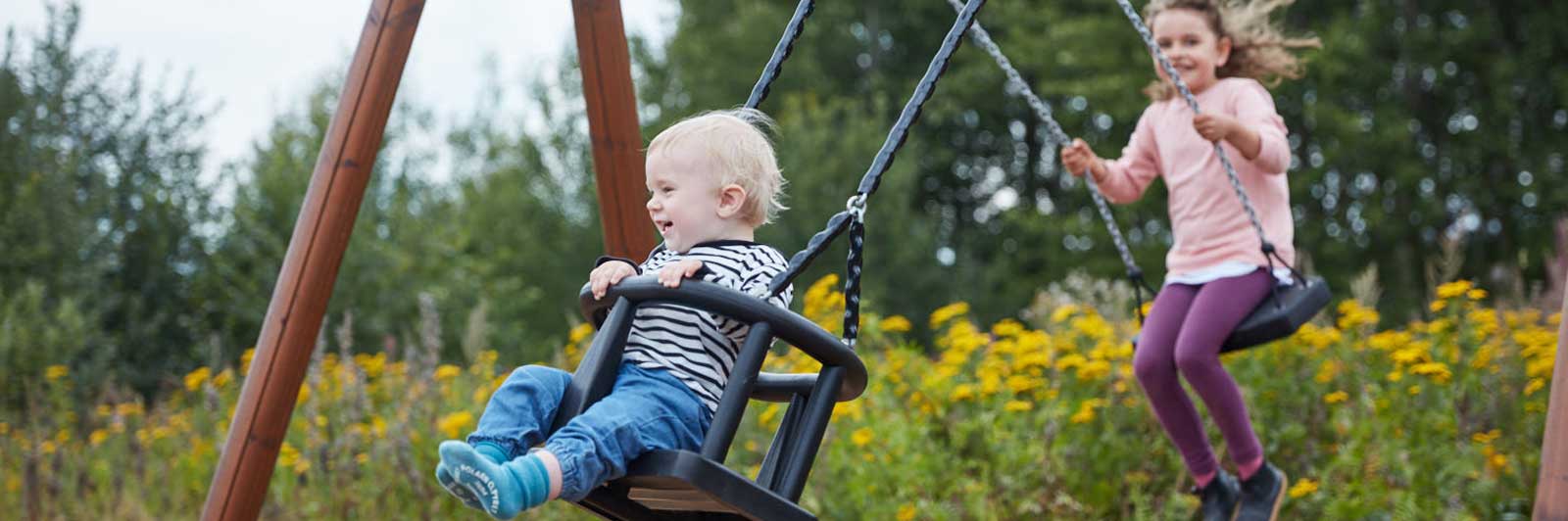 Two children are in the middle of swinging on a swing frame which has 2 swing seats. They are both smiling.